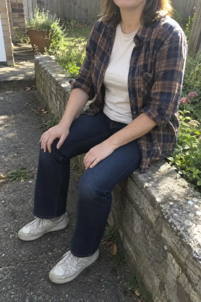 Woman seated on a garden wall wearing an open brown-and-navy plaid shirt, cream tee, dark jeans, and off-white sneakers.