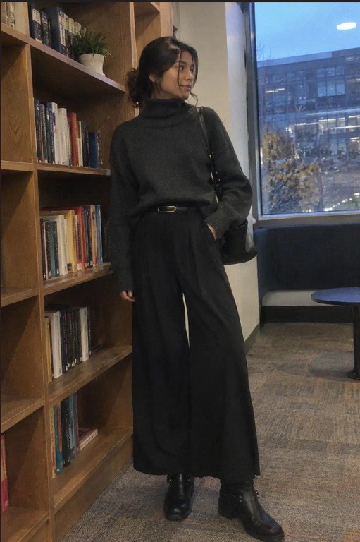 Woman wearing a black turtleneck sweater with high-waisted wide-leg trousers and black leather boots in a cozy office library
