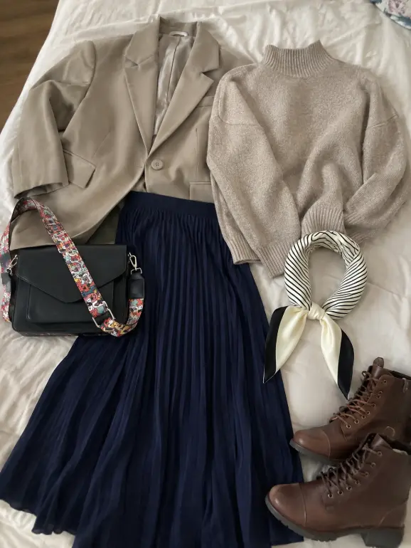 Poetcore outfit flat lay featuring an oatmeal turtleneck sweater, khaki blazer, navy pleated midi skirt, black crossbody bag, striped scarf, and cognac lace-up ankle boots arranged on a white bed.